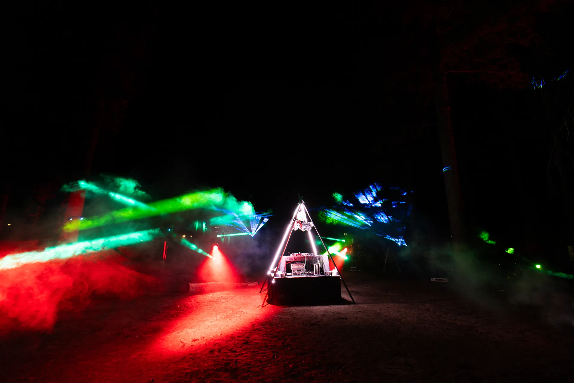 Wide night shot of a triangle DJ booth with red and green lasers cutting through the trees